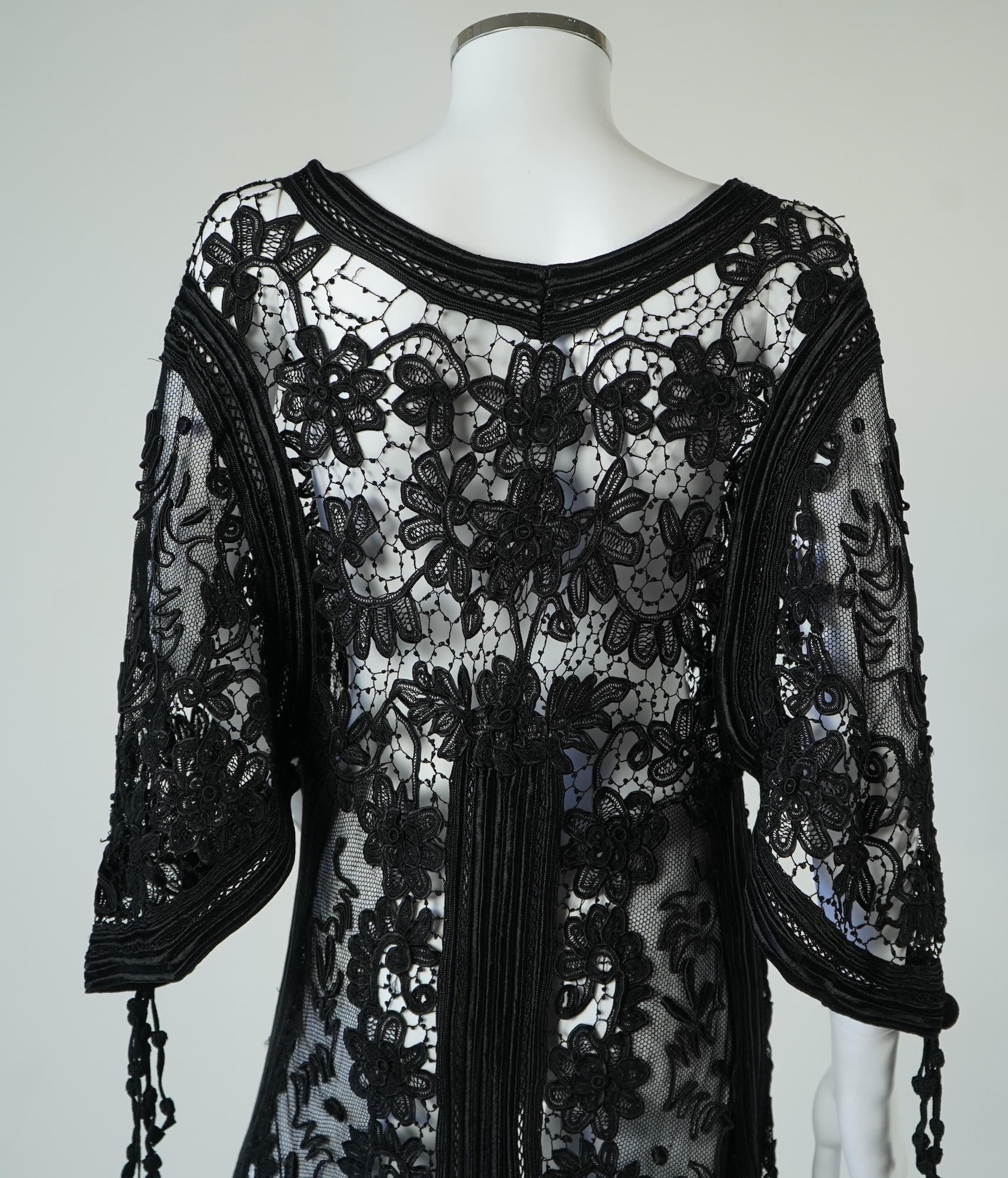A late 19th century stylish ornate black silk tape lace evening coat/dress. Designed with a wide open neck, short bell sleeves with long tassels, four large buttons at the front and tails at the front and back. 130cm lon
